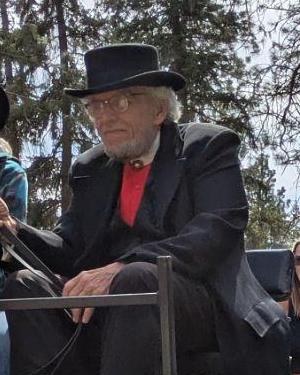 Norman Nelson in a black top hat, seated outdoors among the trees